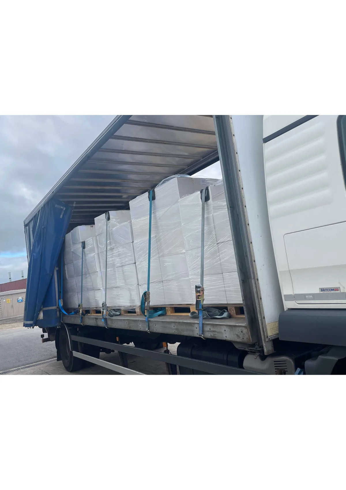 Trailer with curtain side fully open showing strapped shrink wrapped pallets on wooden pallets against a corrugated industrial building during a regional retail goods delivery
